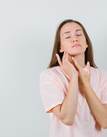 Young woman in pink t-shirt touching skin on neck and chin and looking fascinating , front view.